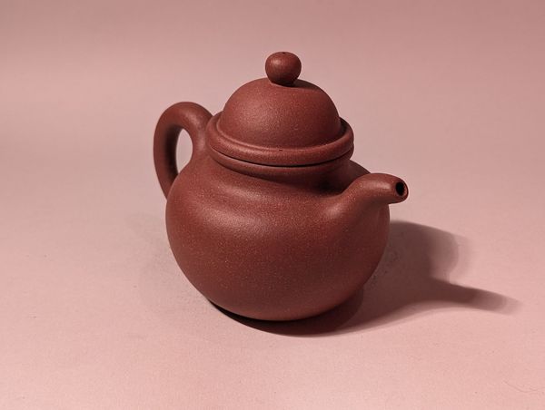 Small brown clay teapot with lid and curved handle on a pink background