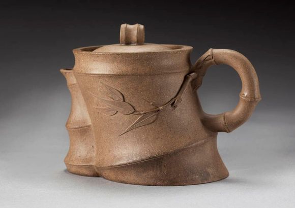 Brown ceramic teapot with a curved handle, lid, and leaf design on a gray background