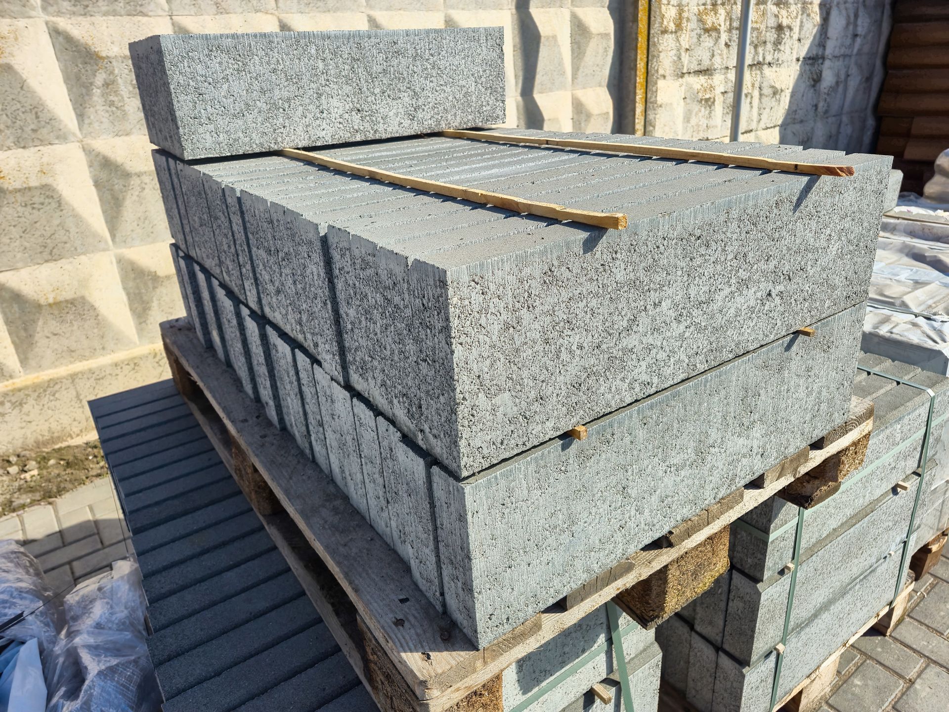 Stacked gray concrete blocks on wooden pallets outdoors beside a textured wall.