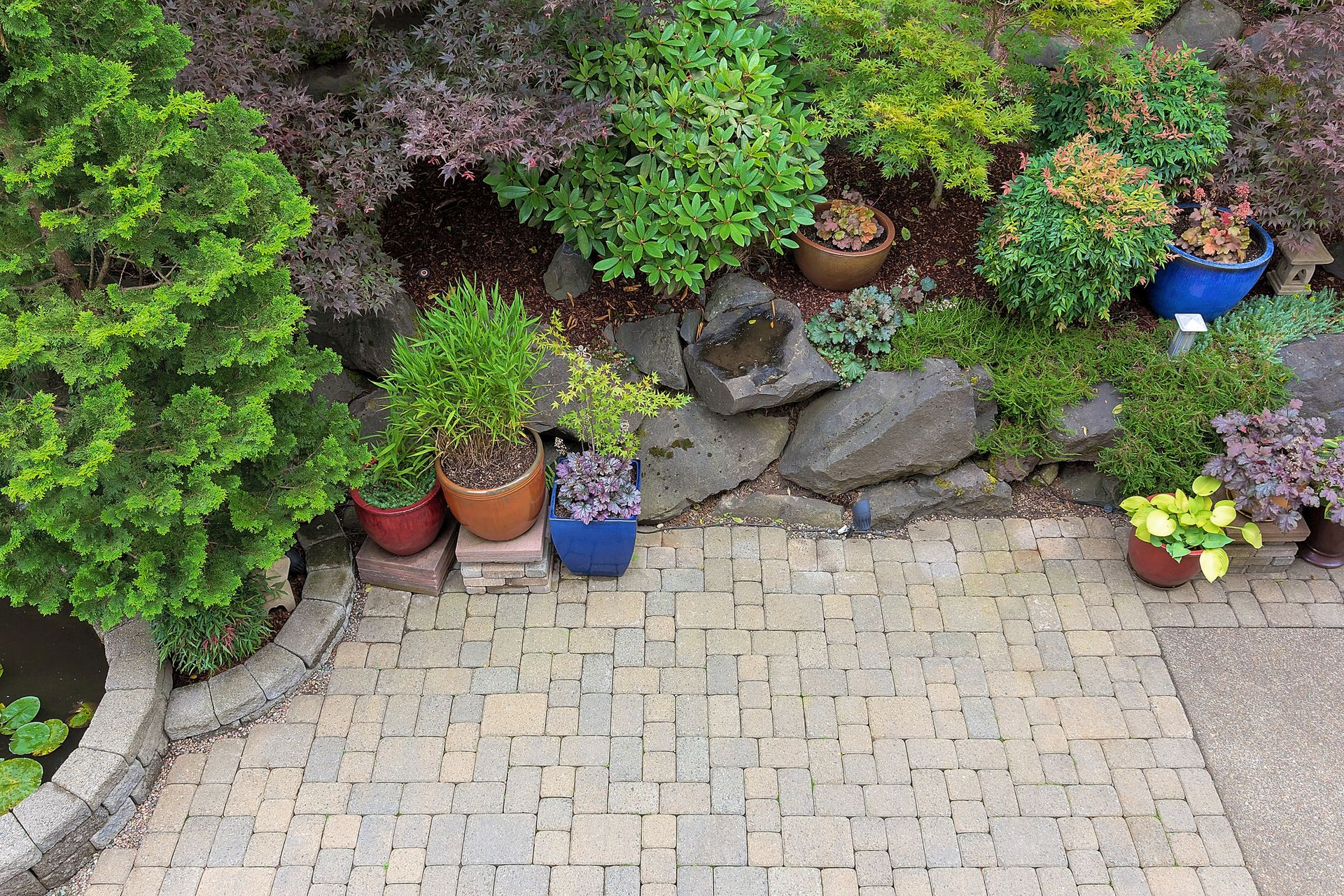 A brick patio framed by a rock garden with various potted plants and lush greenery.