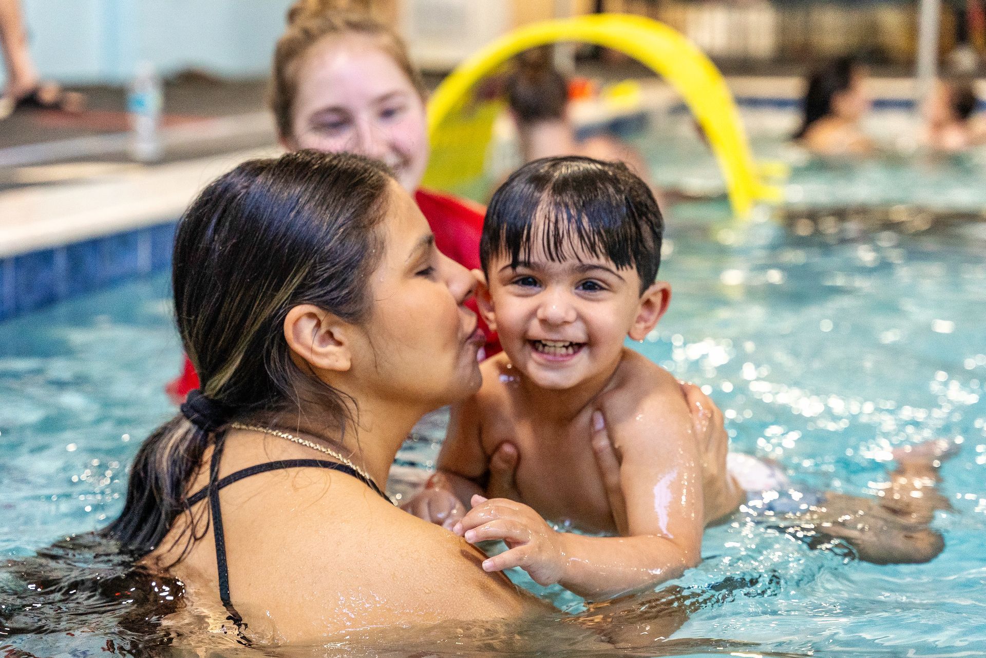 Woman kissing a smiling toddler in a swimming pool. Another person watches.