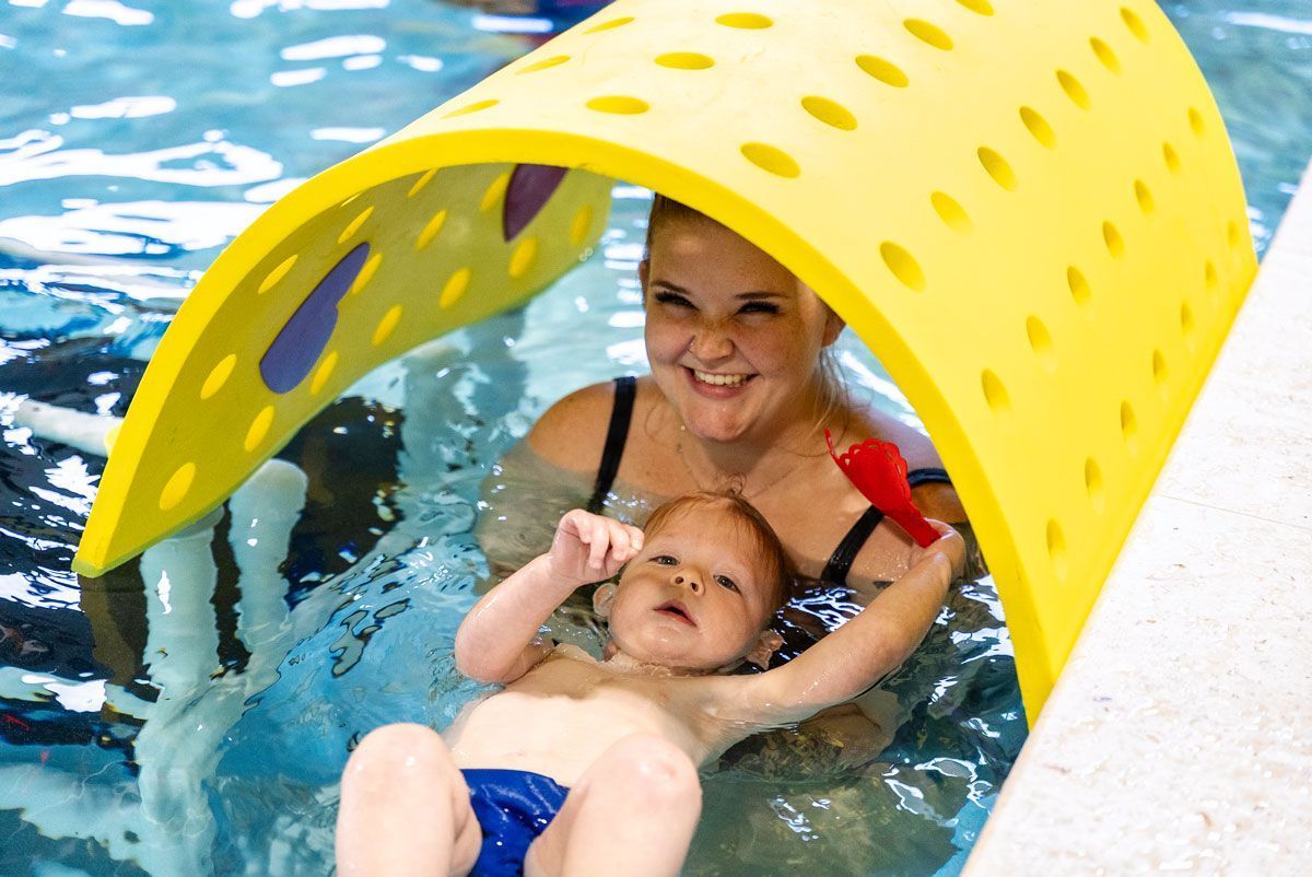 Woman and baby in pool under yellow arch. Woman smiles, baby reaches up, both in water.