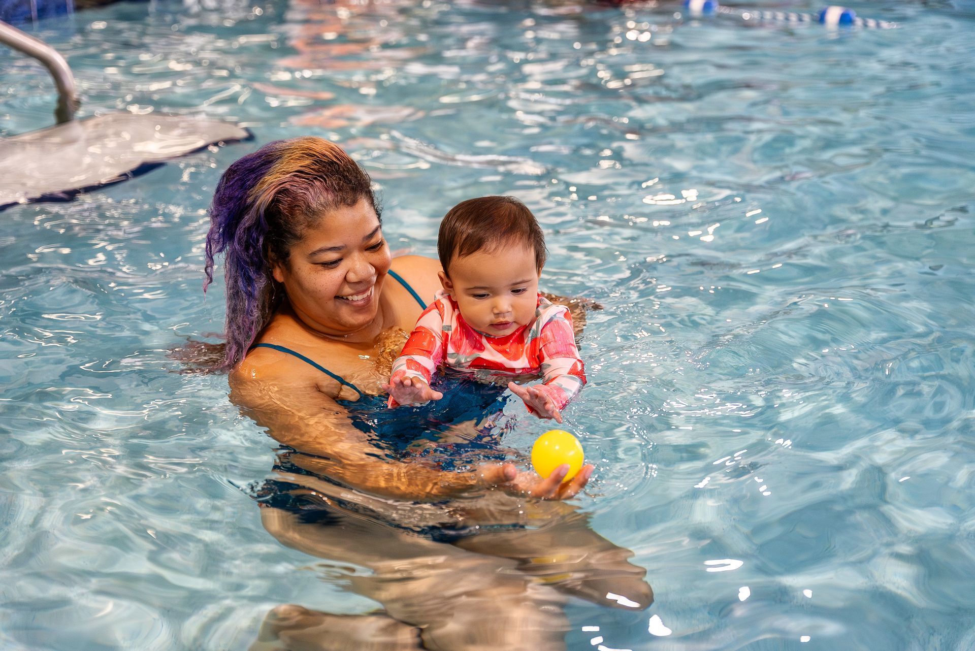 Woman in pool holding baby, both smiling. Baby wearing rash guard, playing with yellow ball.