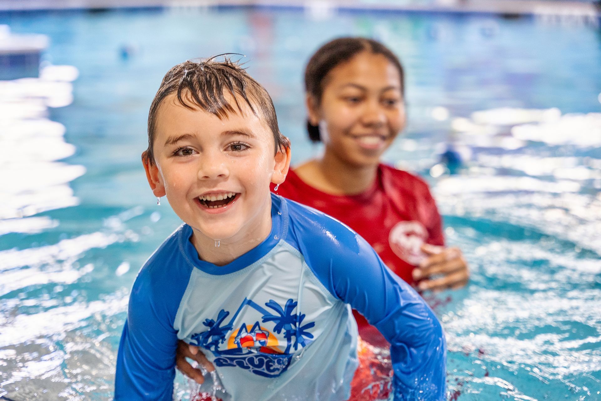 Boy in blue swim shirt smiles in pool with swim instructor behind him.