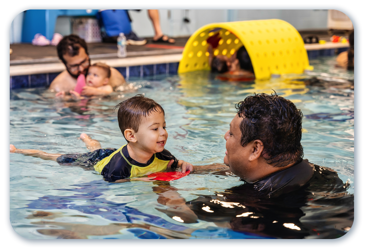 Two children in a pool playing with pool noodles. One child wears a pink cap and goggles; the other wears a blue cap and goggles.
