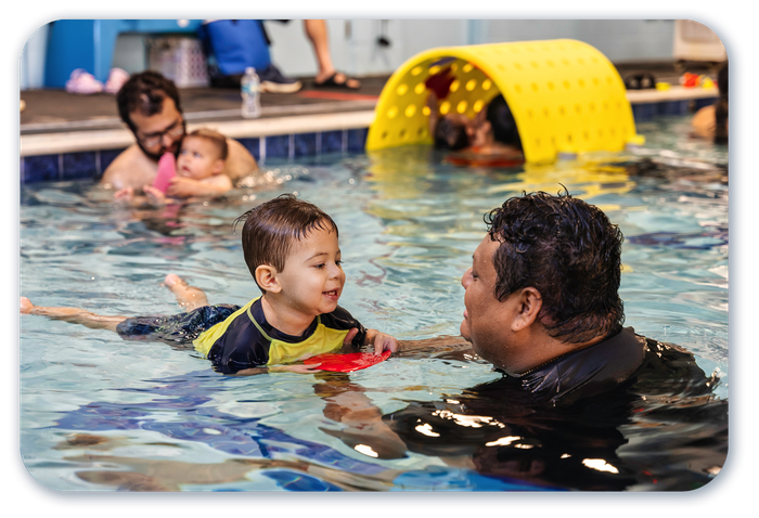 Two children in a pool playing with pool noodles. One child wears a pink cap and goggles; the other wears a blue cap and goggles.
