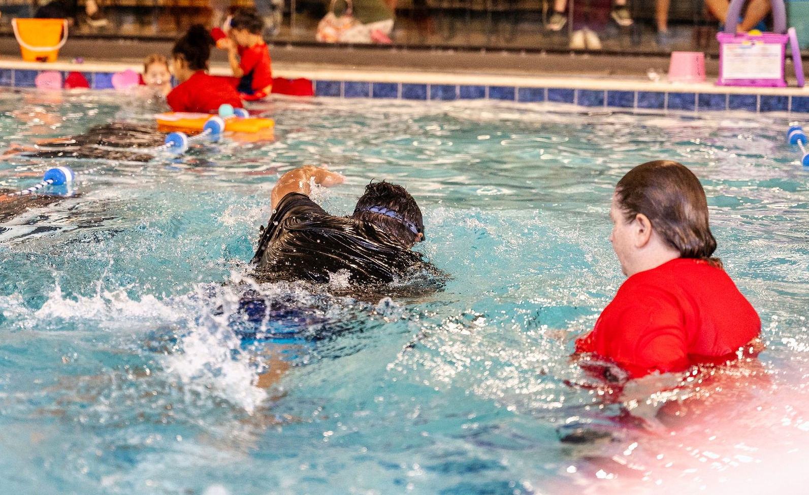 Baby crawls toward camera in shallow pool, swim instructor and other child in background.