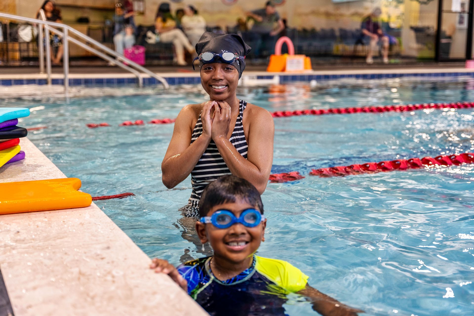 Woman and child in a pool, smiling. Woman wearing a cap and swimsuit. Child in goggles and swimsuit.