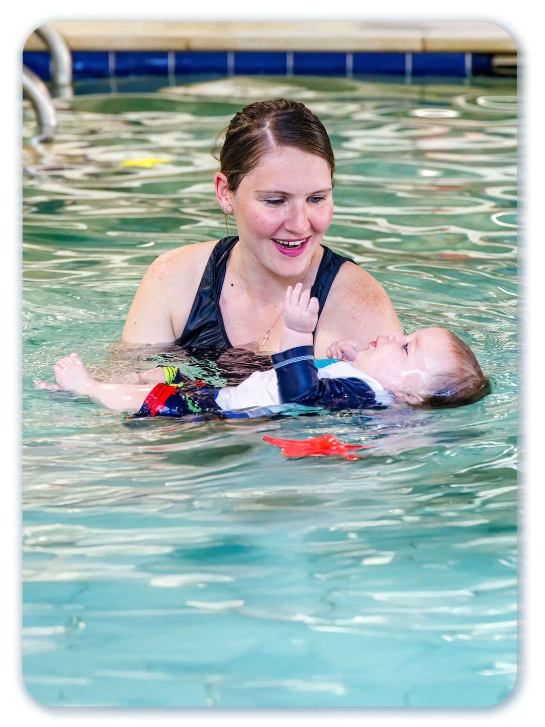 Woman kisses a smiling toddler in a swimming pool with a teacher nearby.