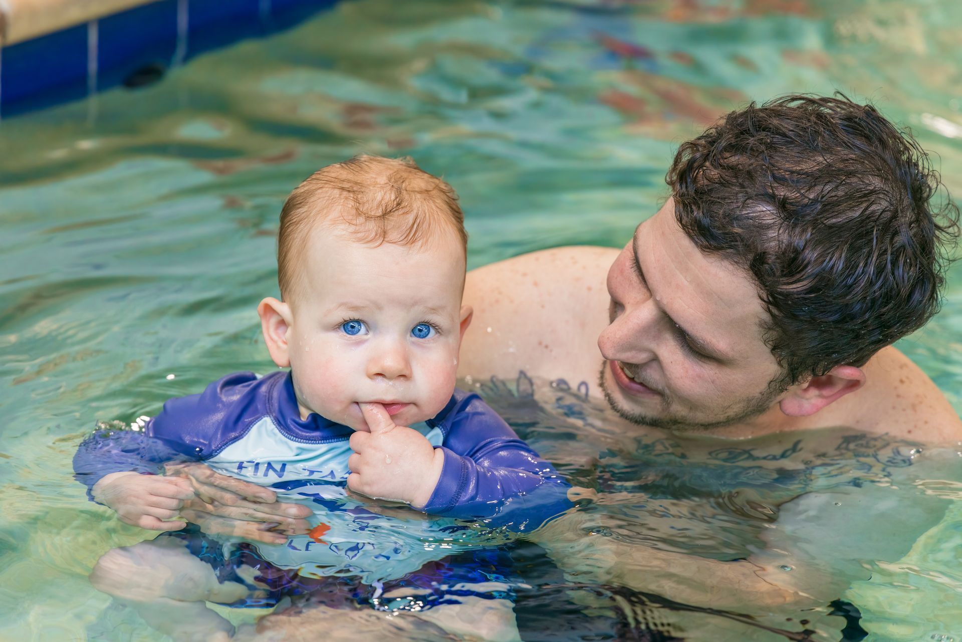 Father and baby in a pool. Baby in blue swim shirt, looks at camera with finger in mouth. Father smiles.