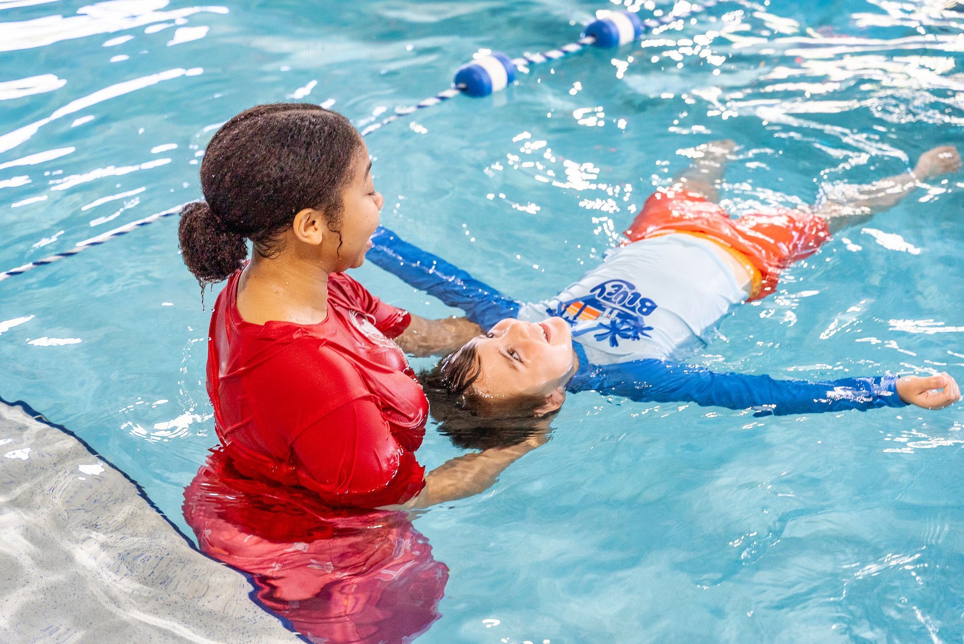 Woman in red shirt teaching a boy to float in a swimming pool; both smiling, clear water.