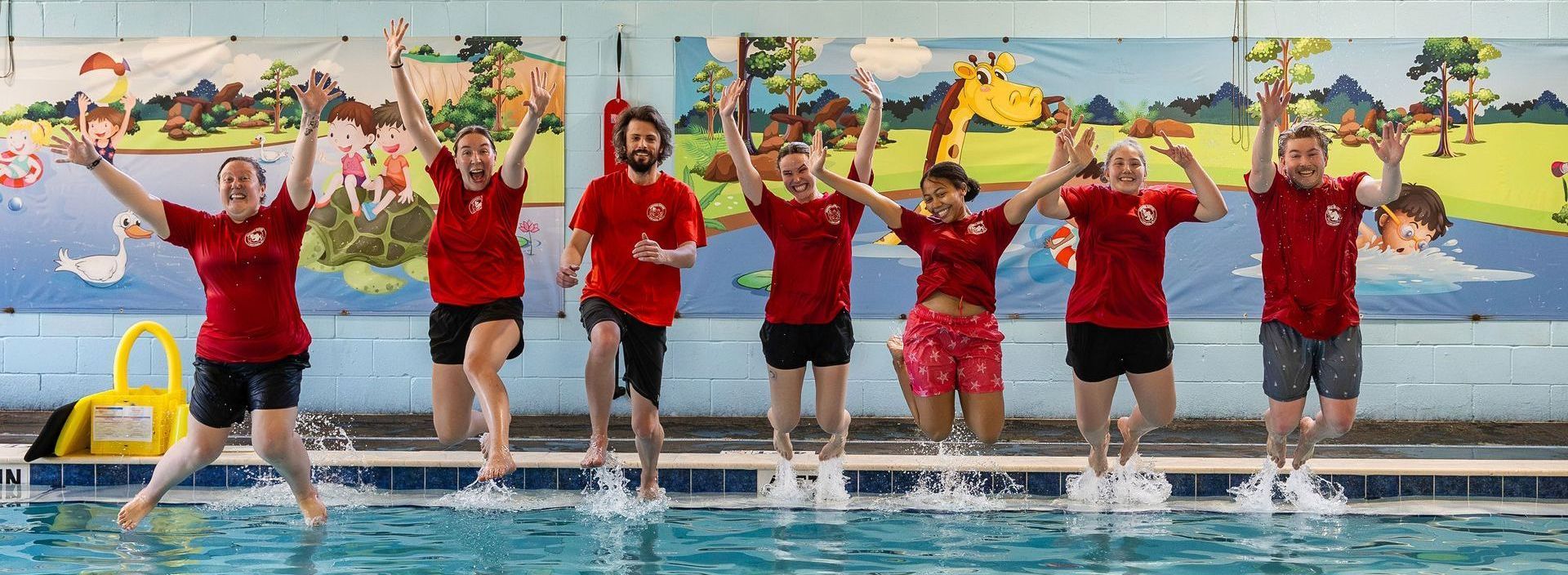 Group of people in red shirts jumping in a pool, hands raised, smiling; colorful mural background.