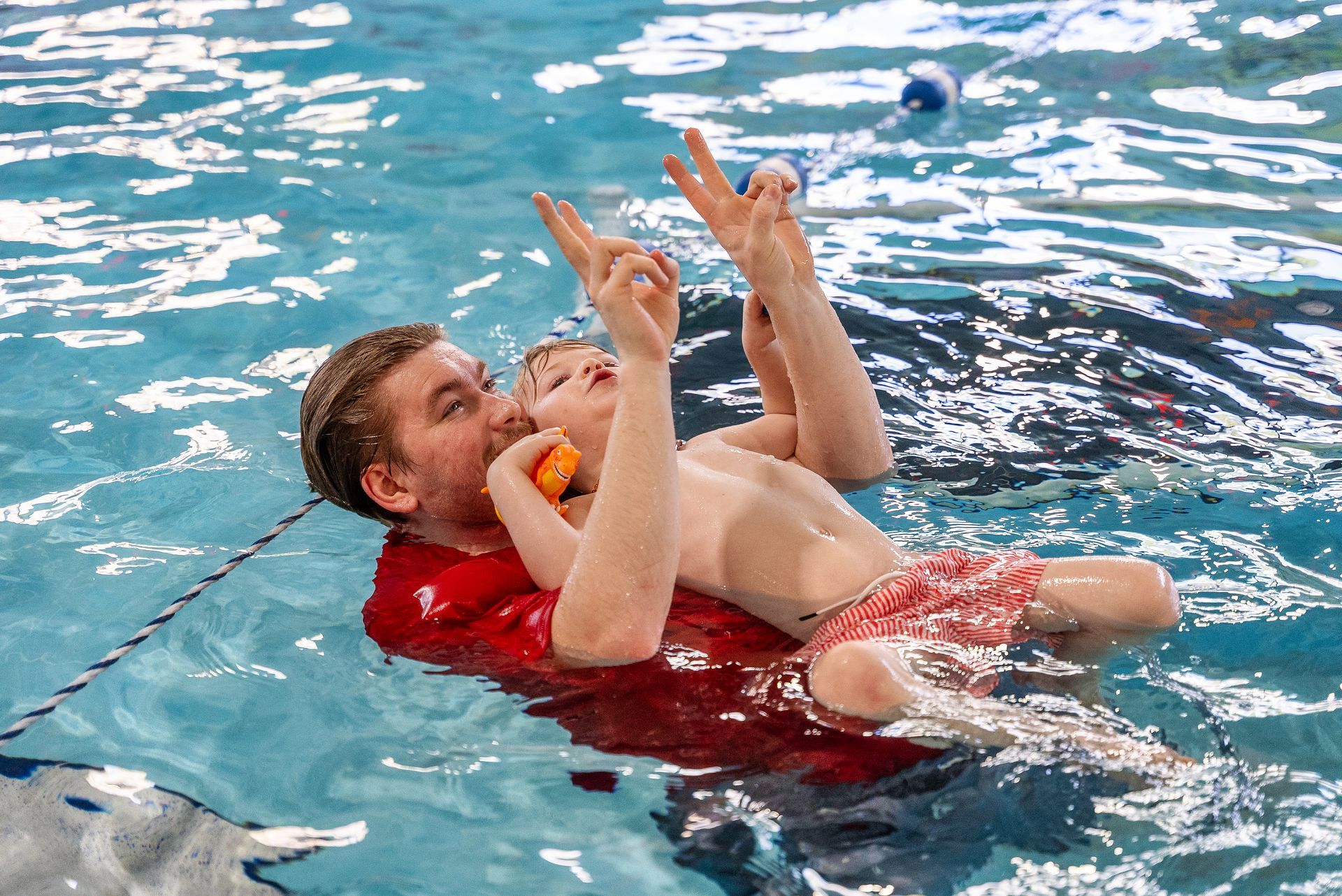 Man in red shirt supports child in a pool, child smiling, making peace signs.
