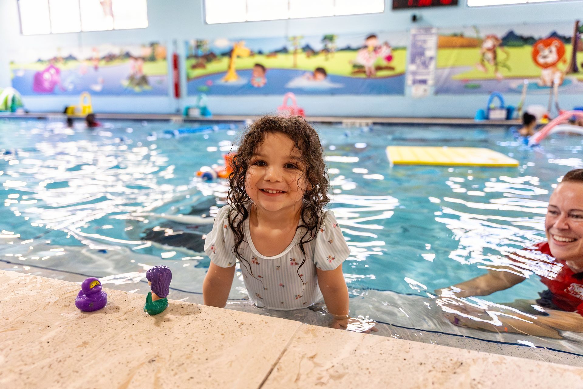 A young girl smiles at the camera while in a swimming pool with a teacher.