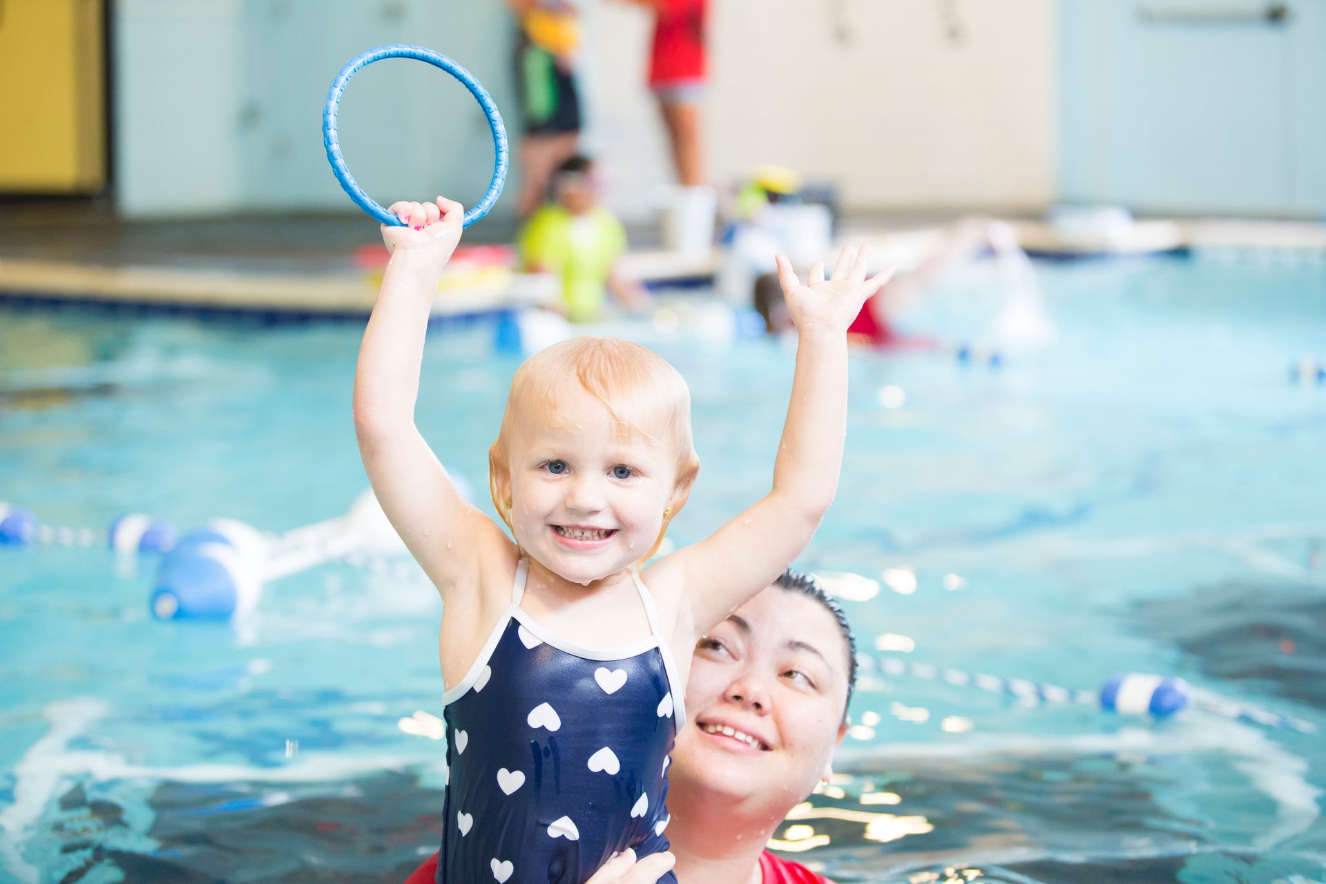 Girl in swimsuit held by woman in pool, holding hoop, smiling.
