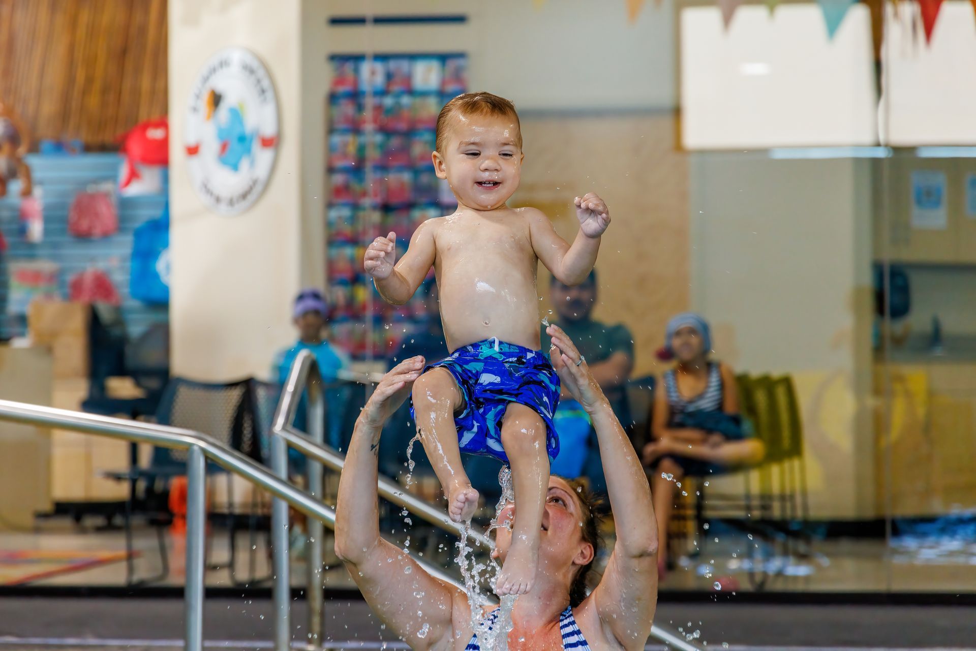Woman holding up a smiling toddler in a pool, splashing water. Other children and a pool setting are visible.