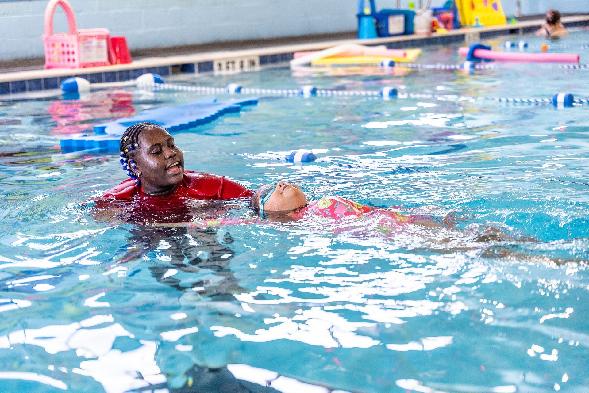 Woman teaching a child to swim in a pool. The woman wears red, the child a swimsuit, both in blue water.
