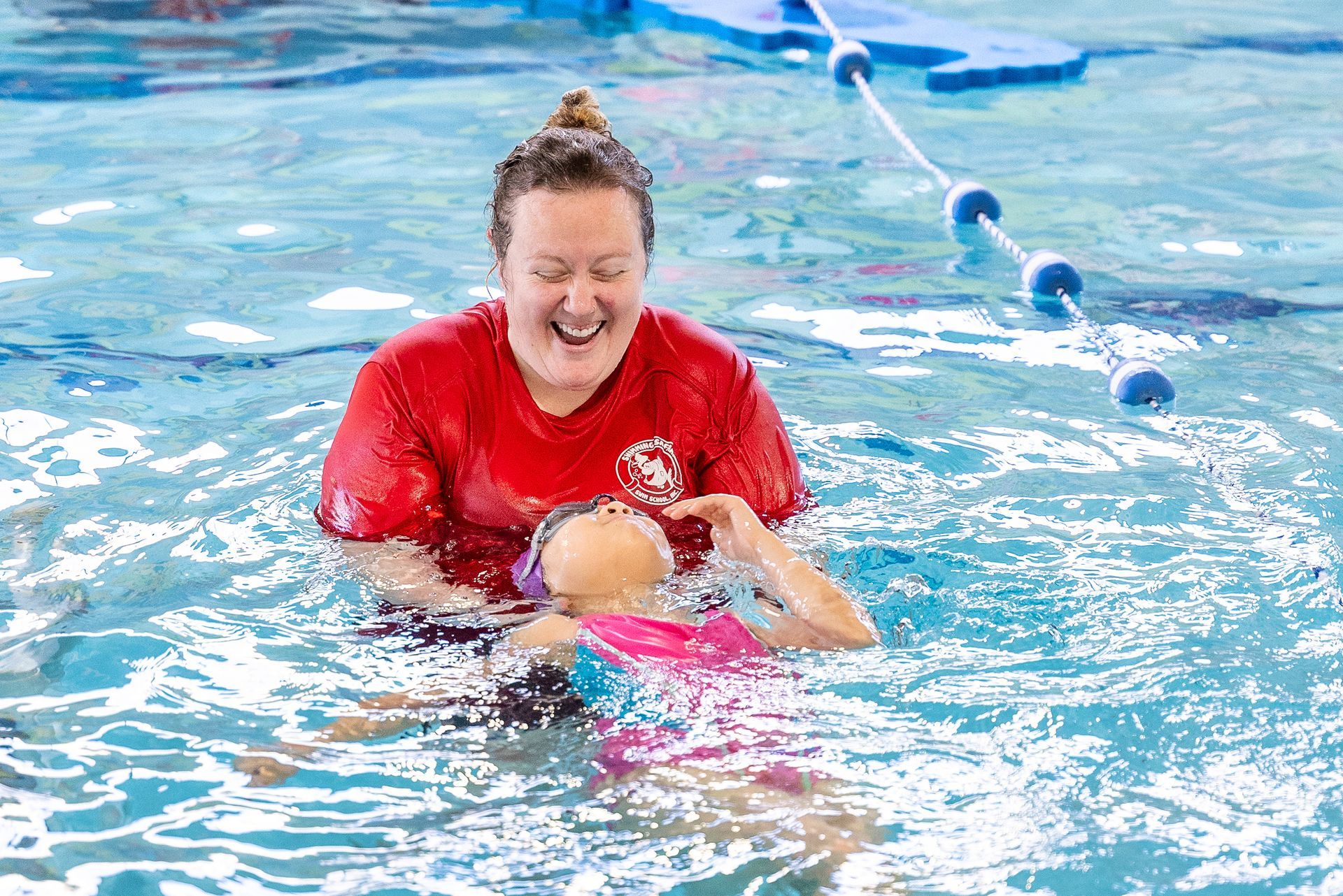 Woman in red shirt teaching a child to swim in a pool. They are smiling in the water.