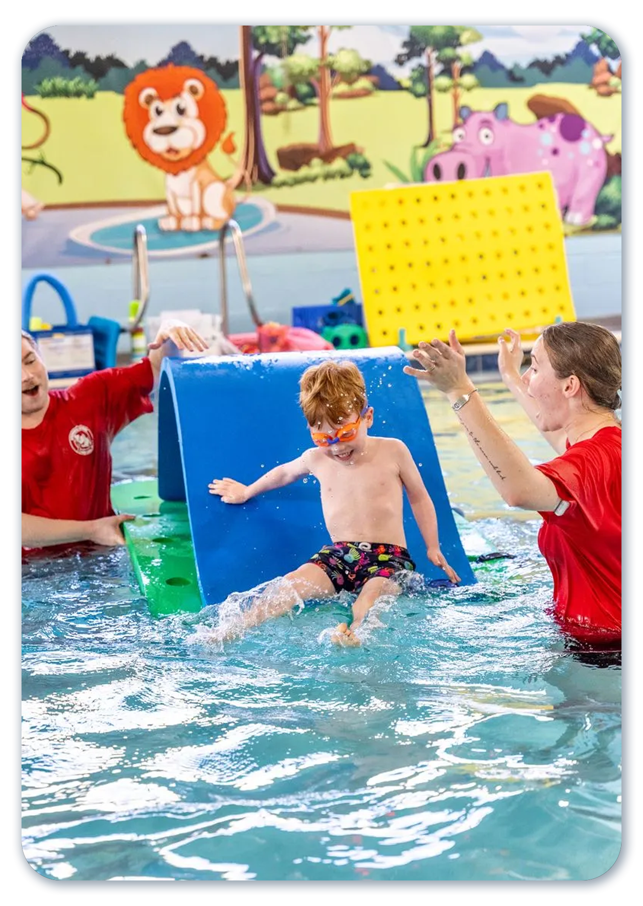 Boy slides into pool from a blue ramp, guided by two adults in red shirts, in a swim class setting.