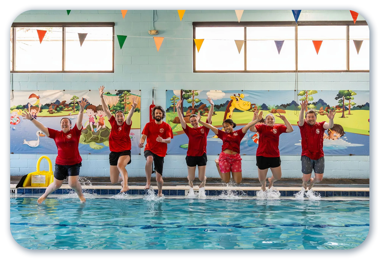 People in red shirts jumping in a pool, arms raised. Poolside, colorful backdrop and decorations.