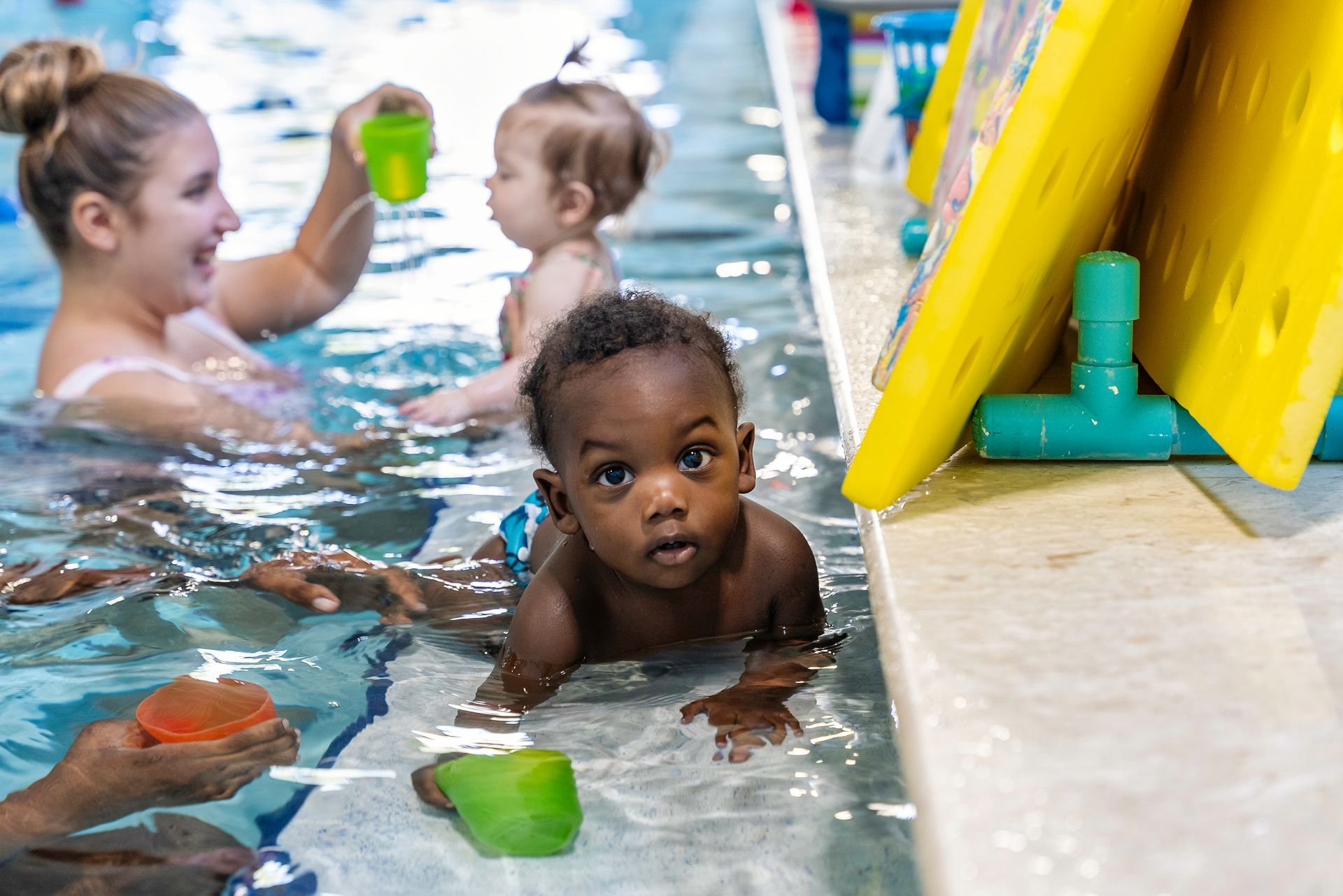 Baby crawls toward camera in shallow pool, swim instructor and other child in background.