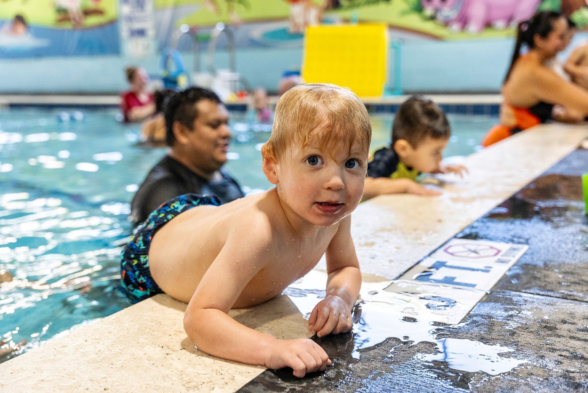 A young child with wet hair smiles at the camera while holding onto the edge of a swimming pool.