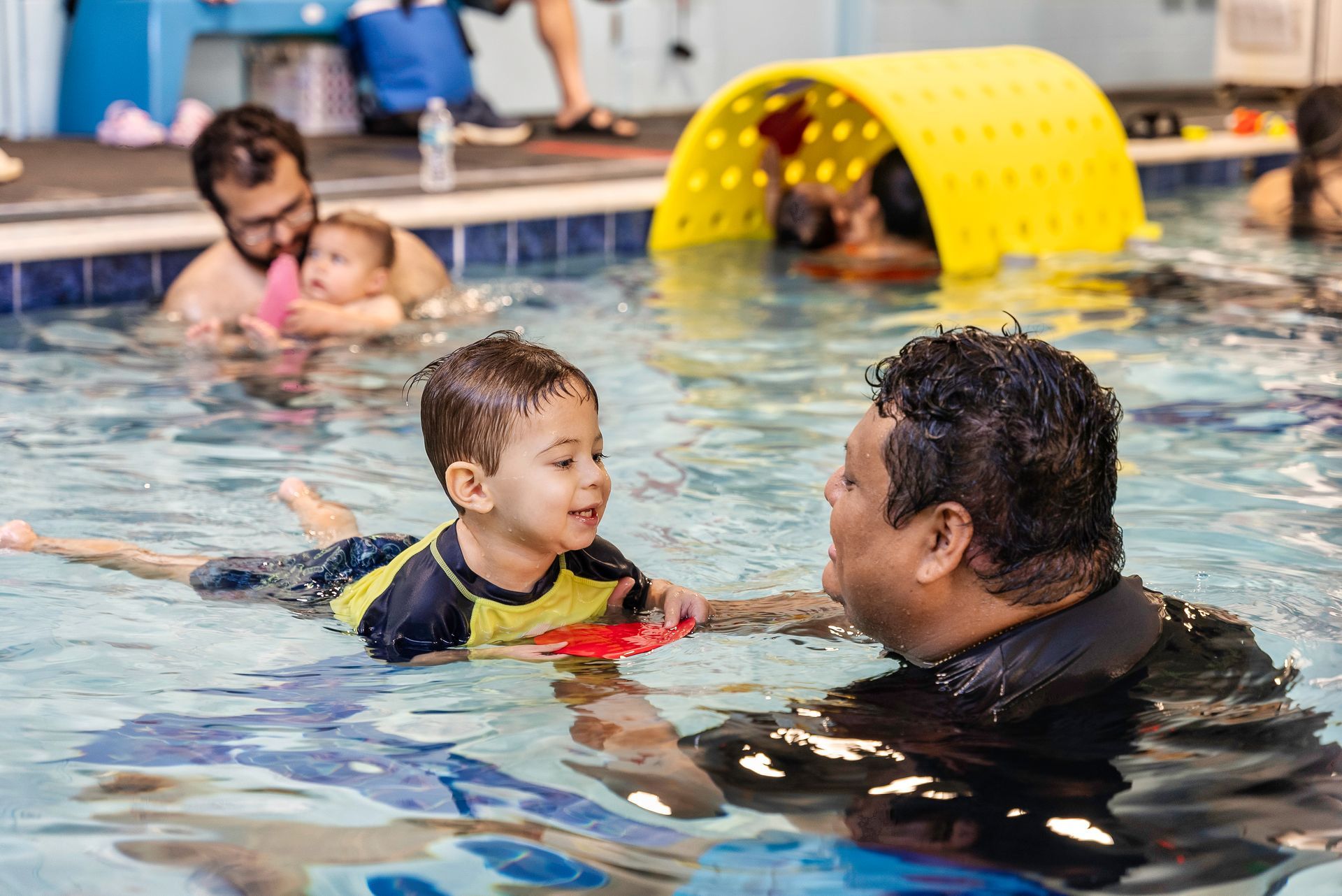 Child learning to swim with an instructor in a pool; other parents and children also present.