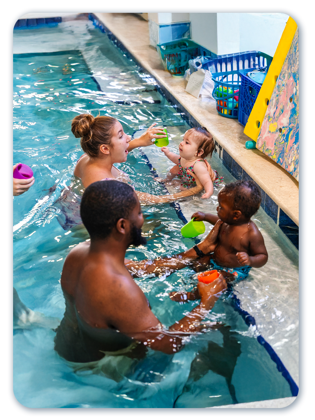 Adults and babies playing in a pool. One adult hands a toy to a baby. Sunlight reflects on the water.