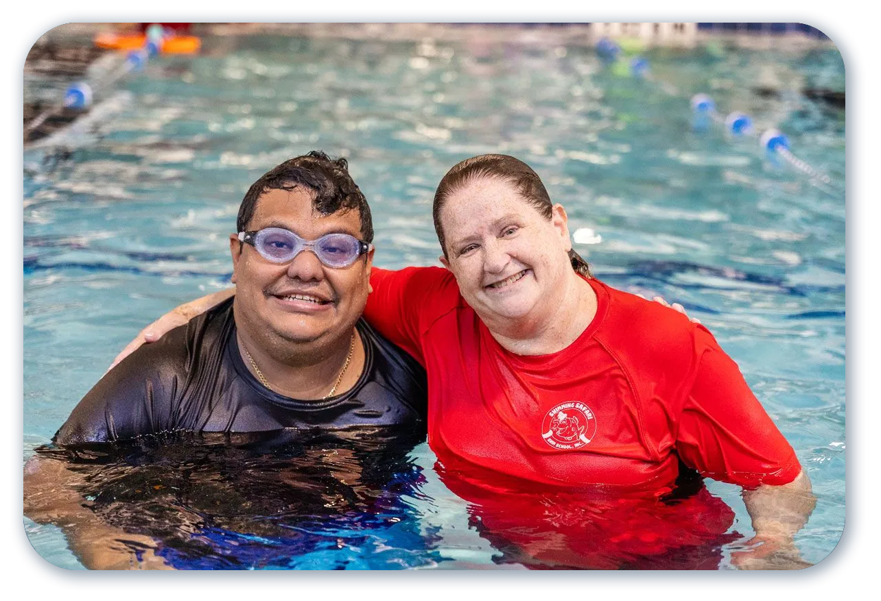 Two people smiling in a swimming pool. One has goggles. One wears a red shirt and has arm around the other.