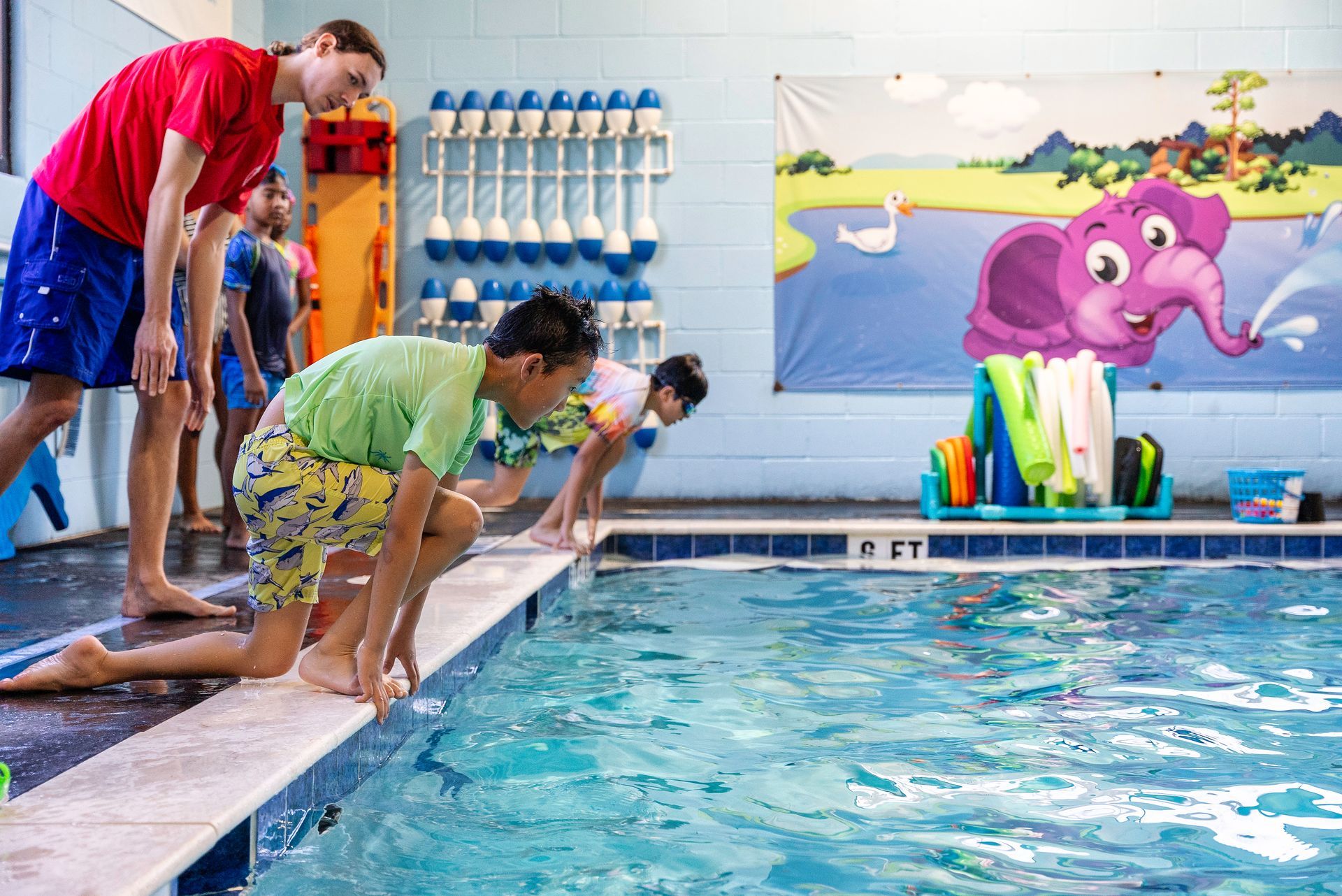 Children in swimsuits preparing to jump into pool, supervised by an instructor.
