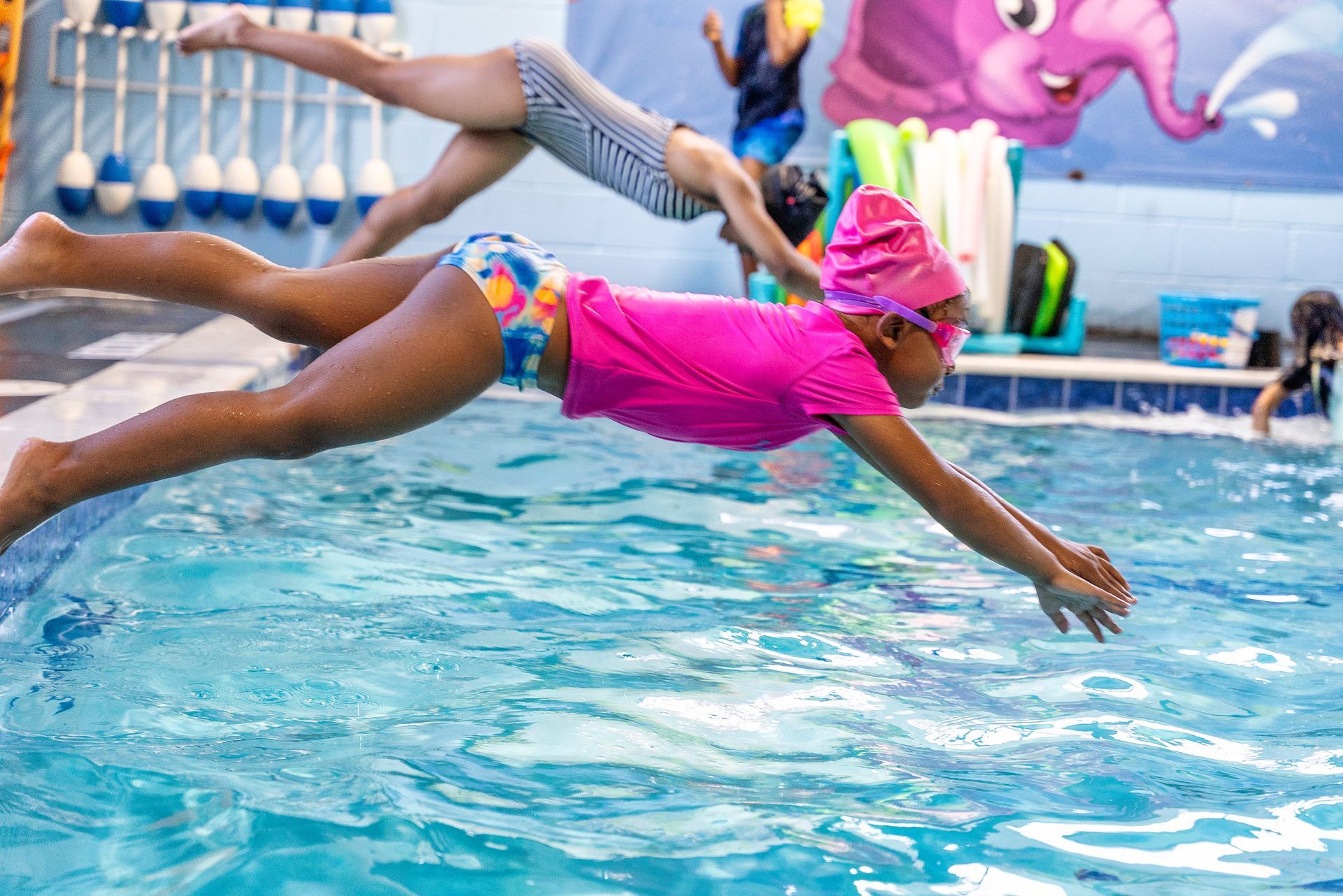 Girl in pink shirt diving into pool; another in background. Indoor pool setting.
