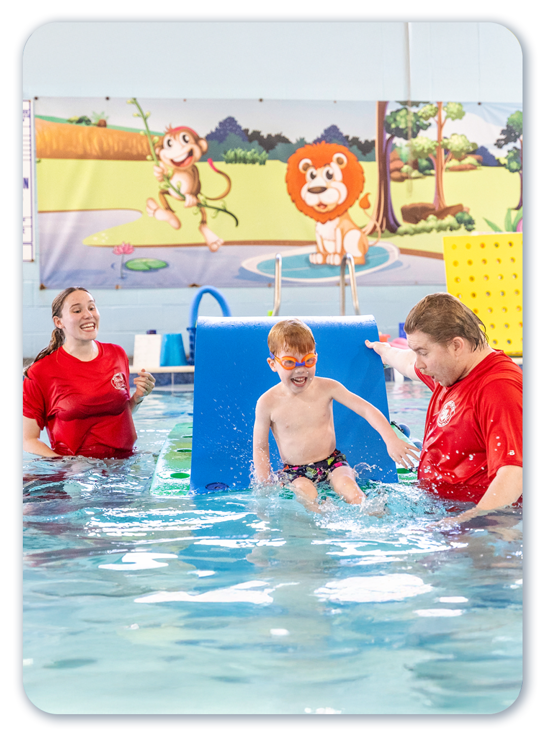 Child in swim class, splashing between instructors. Pool setting with jungle mural.