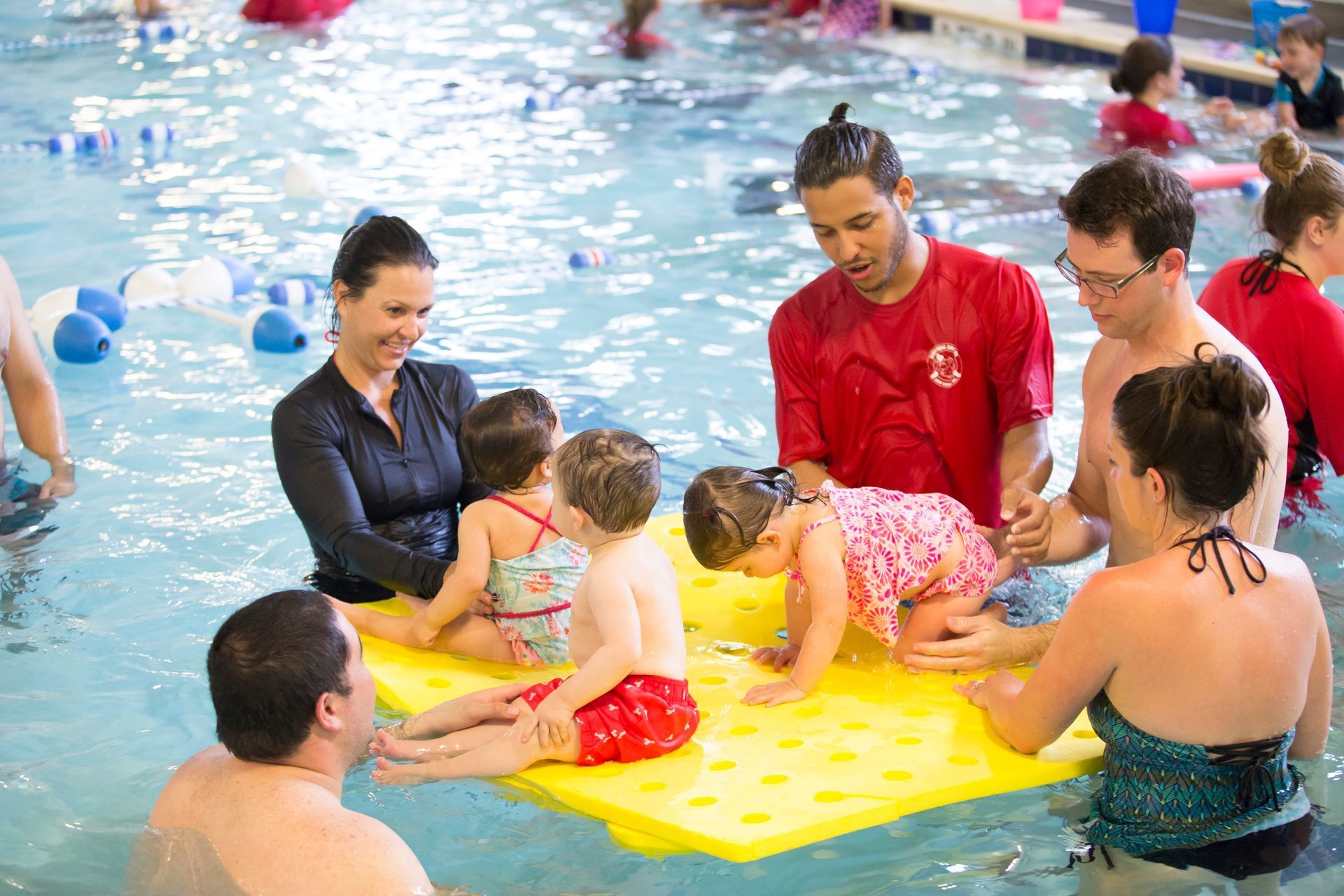 Parents and instructors in a swimming pool with toddlers on a yellow floating mat.