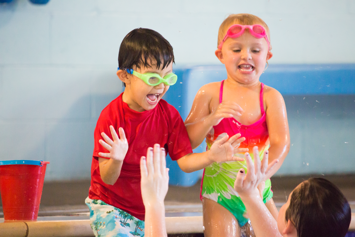 Two children in swimwear at a pool are splashing and laughing. A woman in the water plays with them.