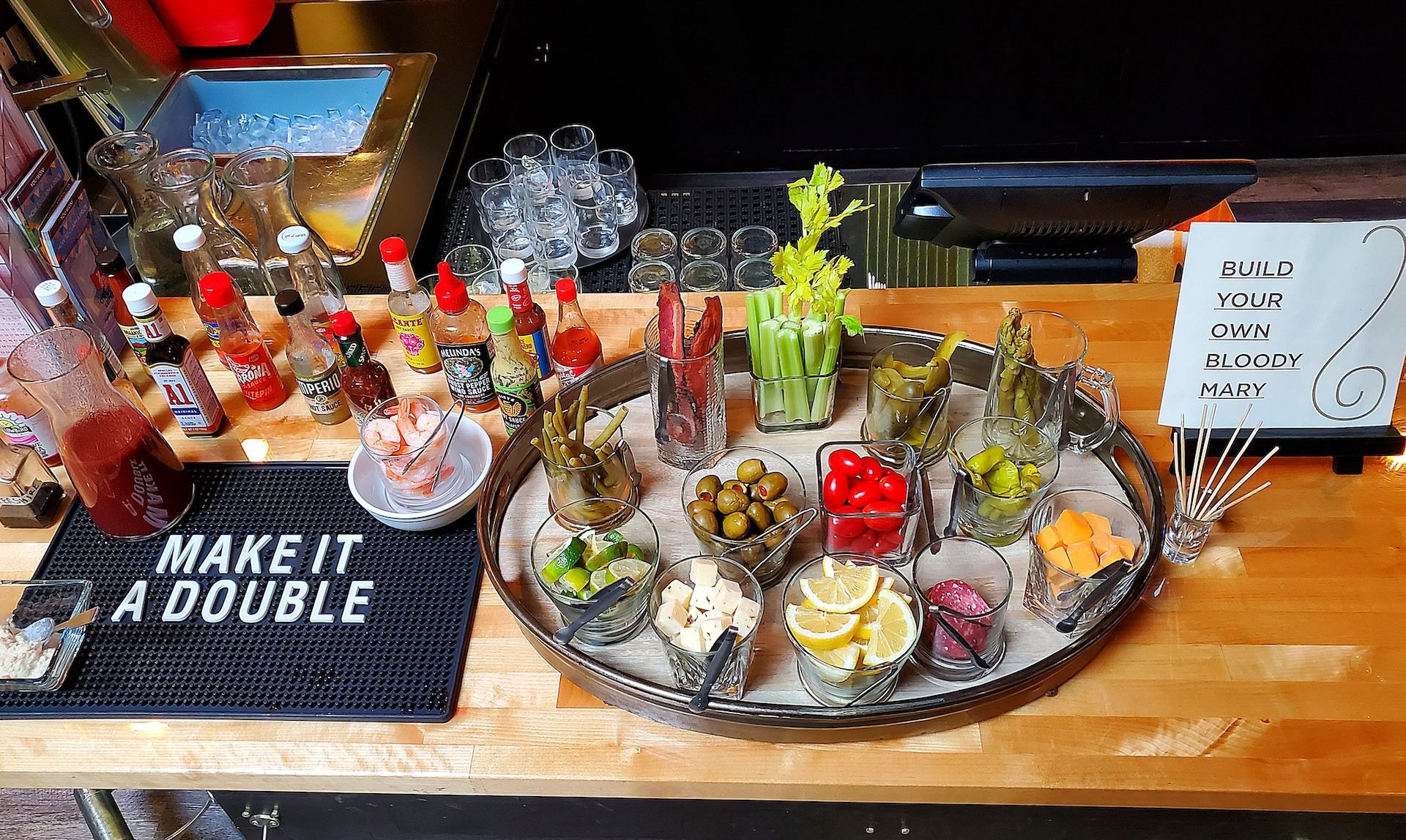 A cocktail garnish tray with various ingredients on a wooden bar, plus bottles and a sign that says 