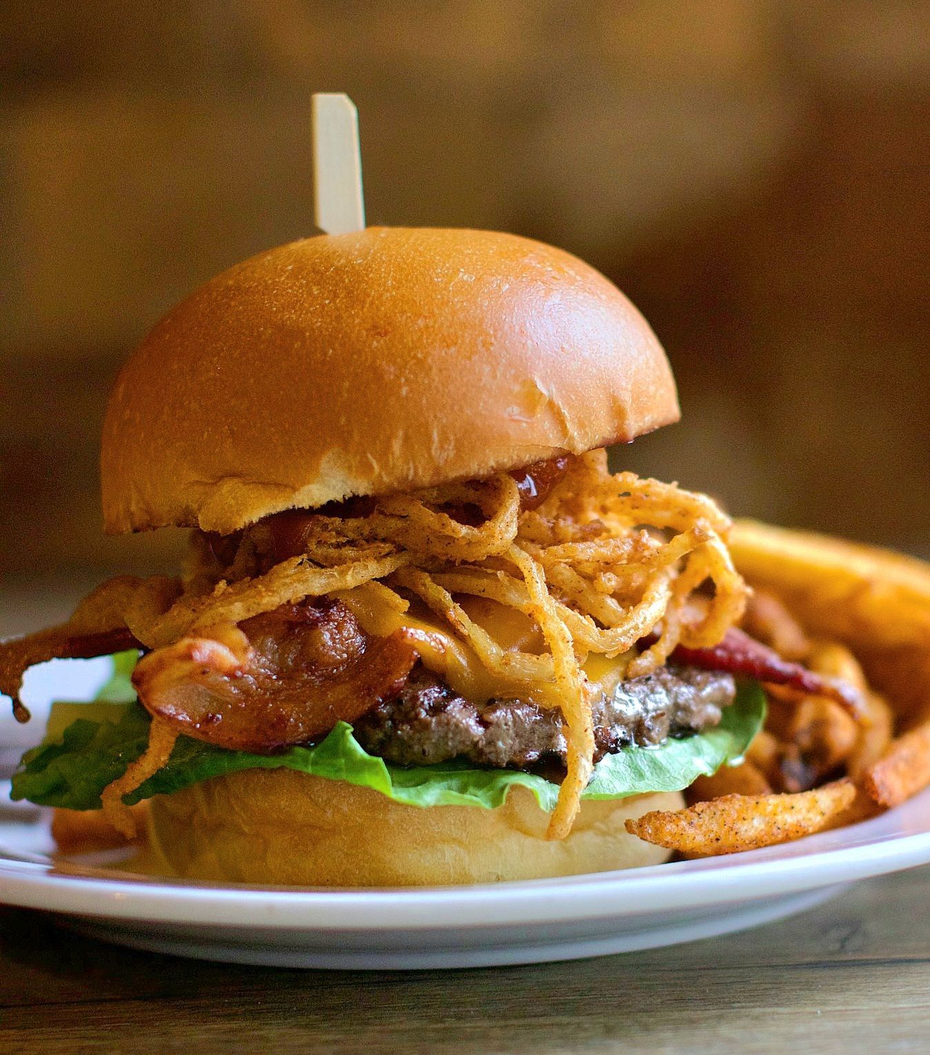 Burger with lettuce, bacon, onion rings, and a wooden skewer on a white plate.