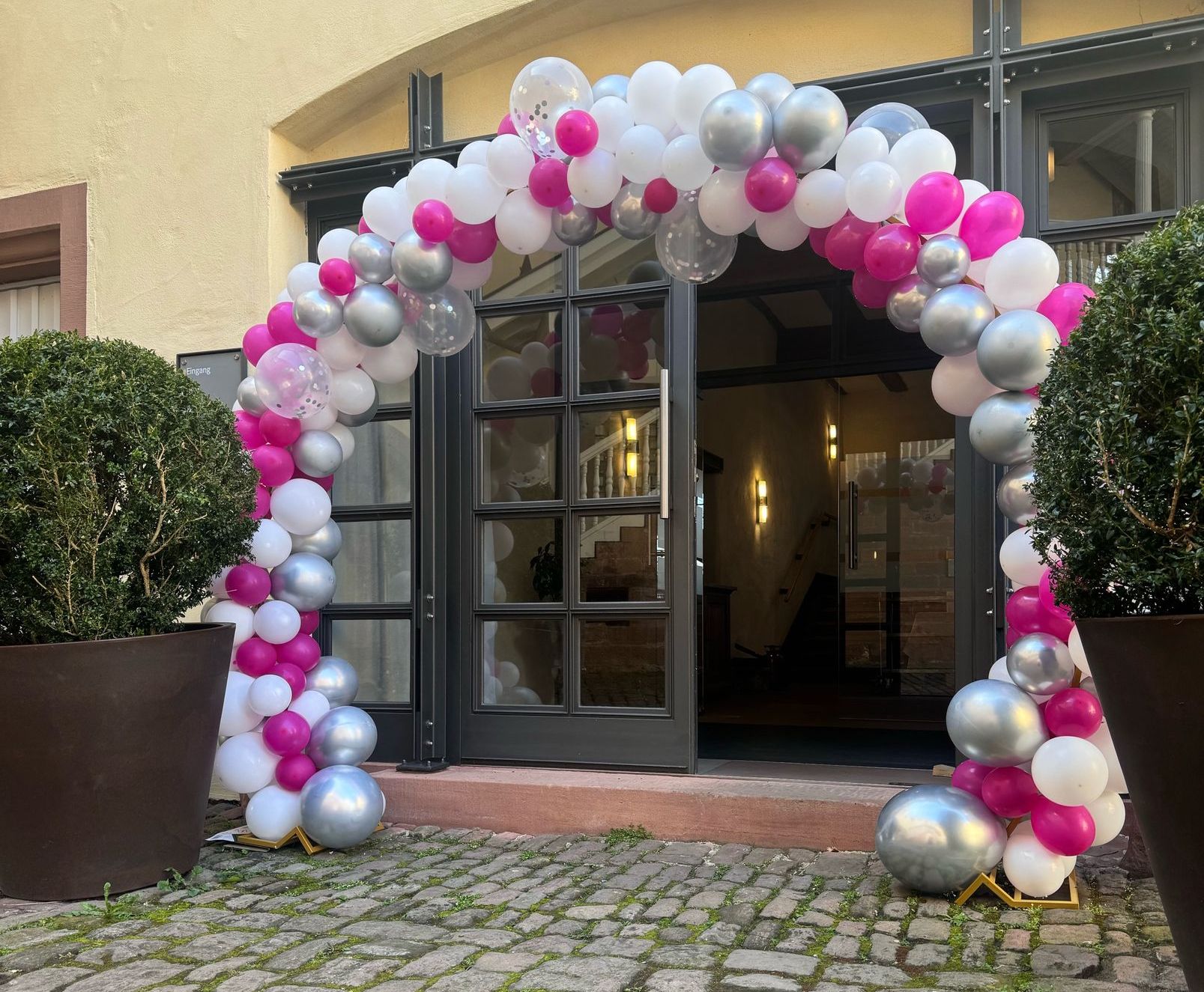 Ein Ballonbogen in Weiß, Rosa und Silber schmückt einen Türrahmen.