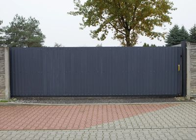 A black gate is sitting on a brick driveway next to a tree.