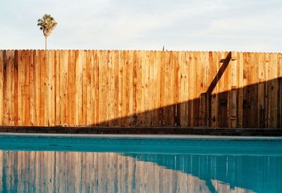 A wooden fence surrounds a swimming pool with a palm tree in the background.