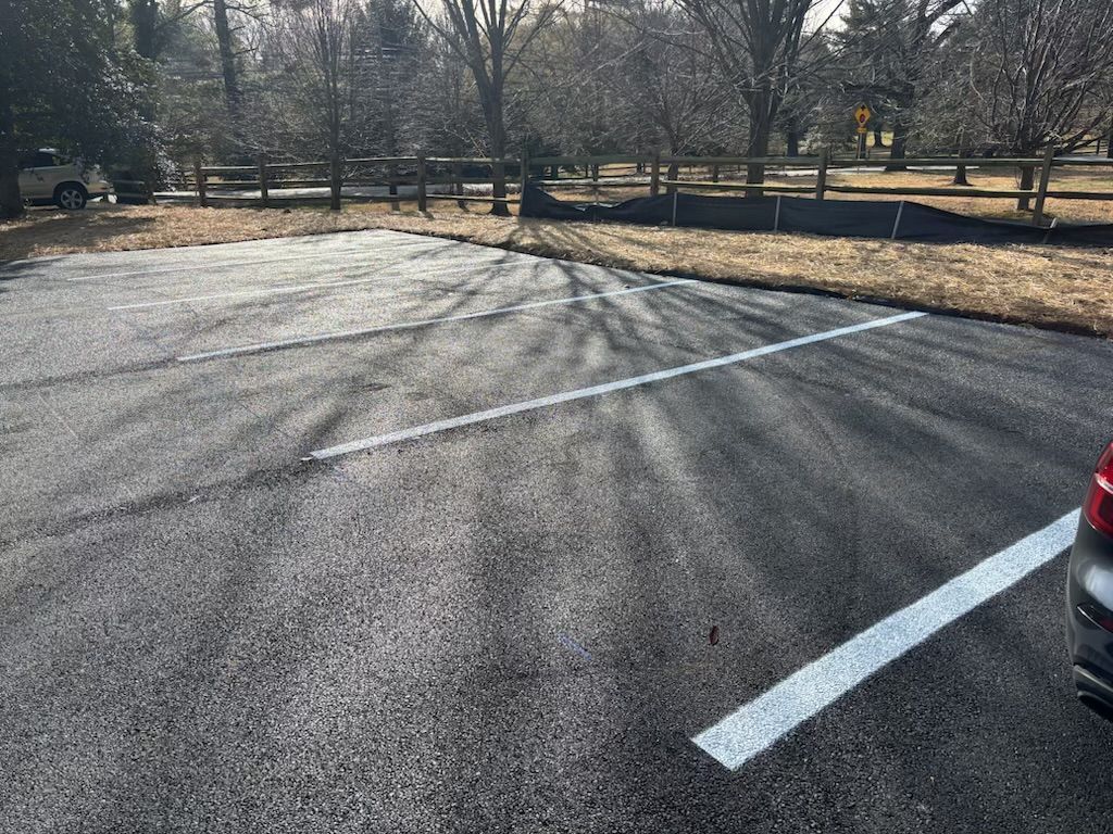 A paved parking lot with white painted stall lines, wooden perimeter fencing, and trees in the background.
