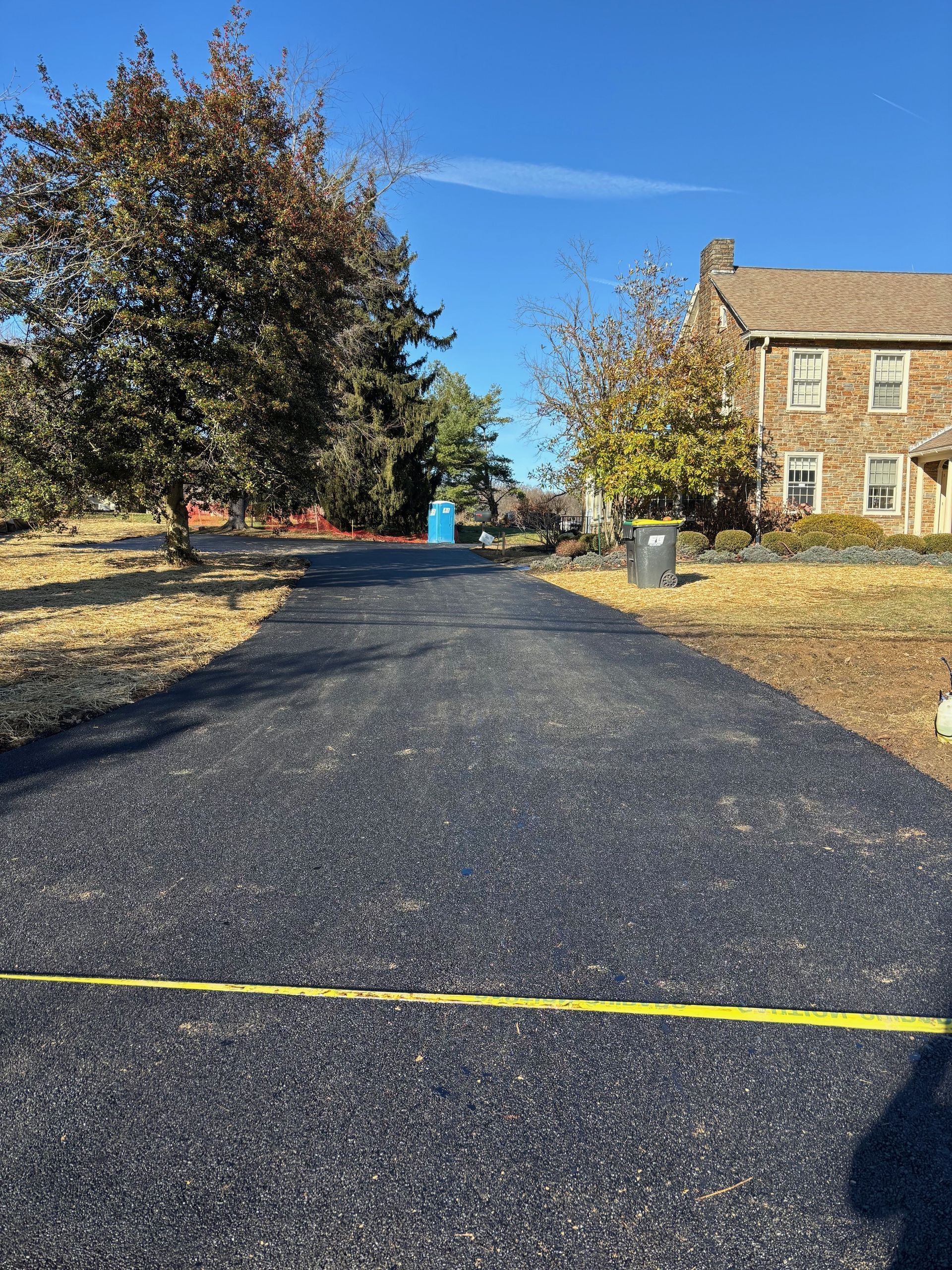A newly paved asphalt driveway leads toward a stone house on a sunny day, with a yellow tape line across the foreground.
