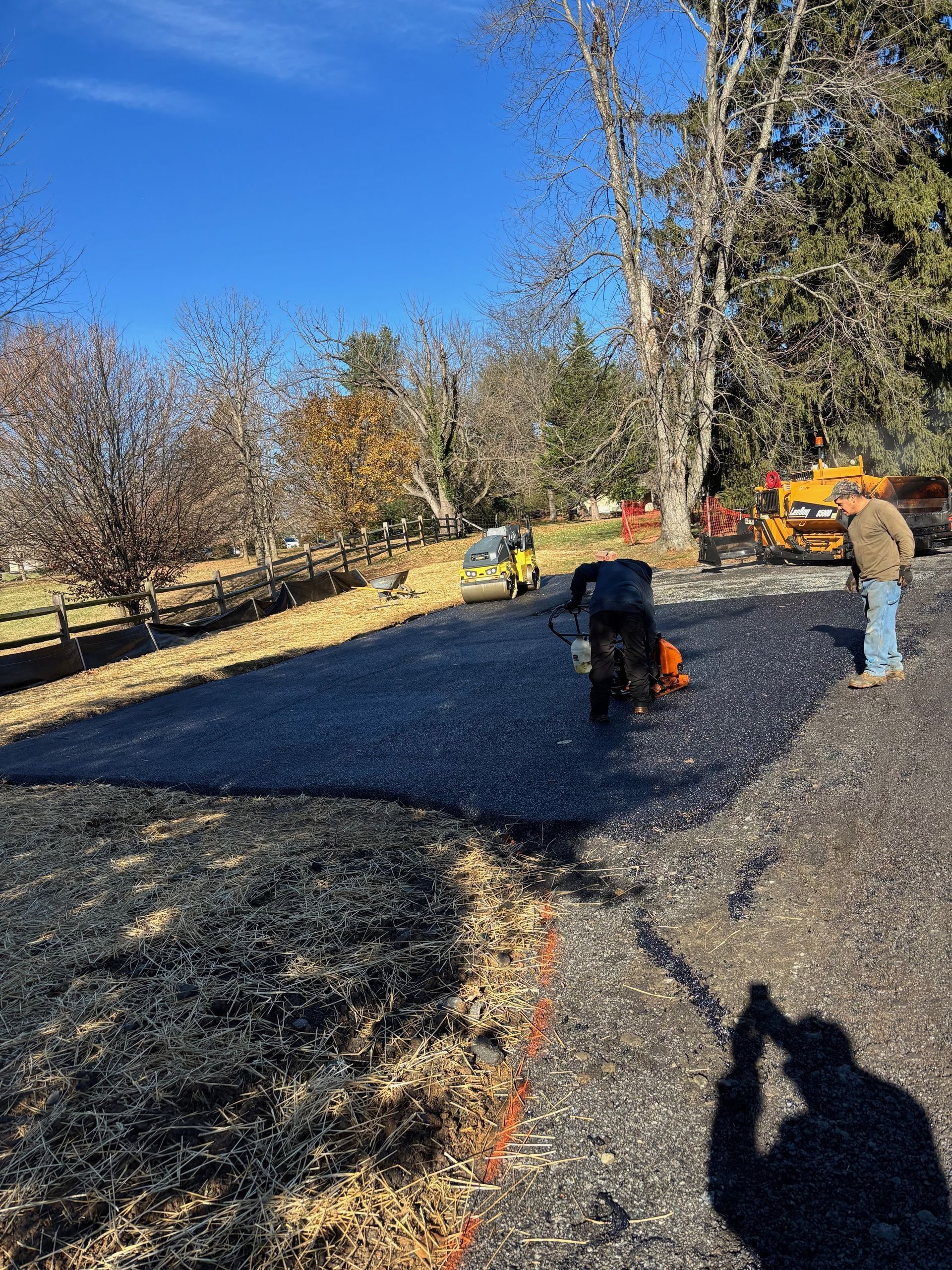 Two workers pave a driveway with black asphalt under a bright blue sky, with a small steamroller and truck nearby.