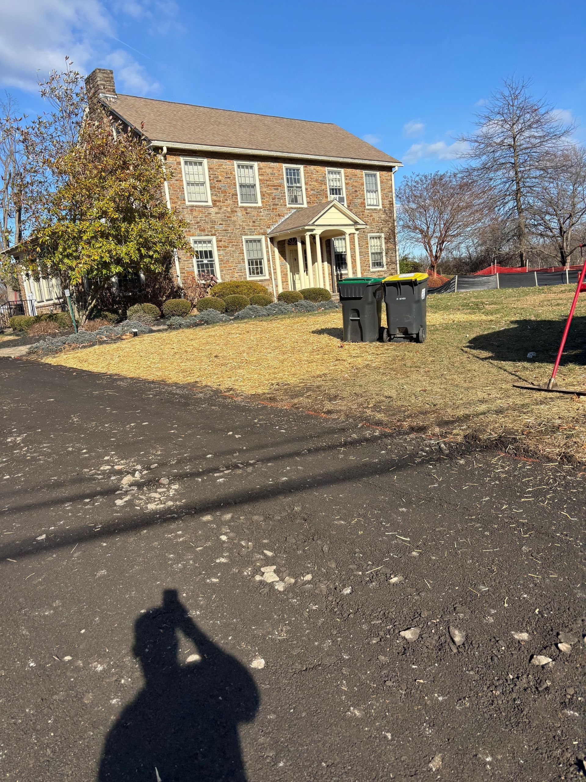 A two-story stone house with a shingled roof stands behind a lawn with two trash bins, under a clear blue sky.