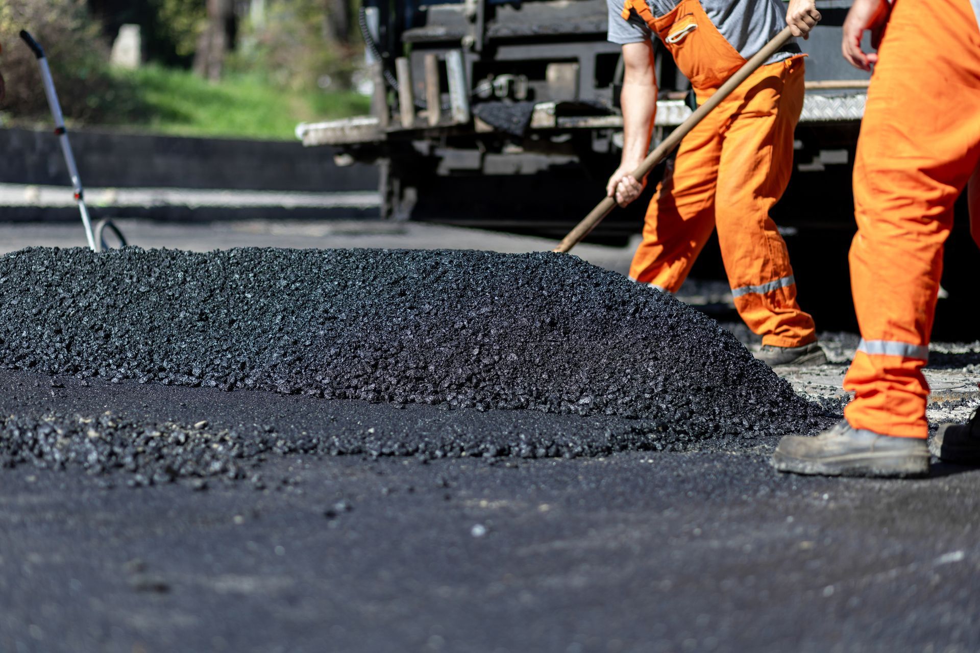 Two workers in orange uniforms spread asphalt on a road.