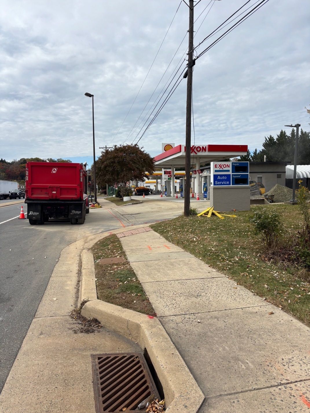 Concrete sidewalk with a drain, a red dump truck, and a gas station under an overcast sky.