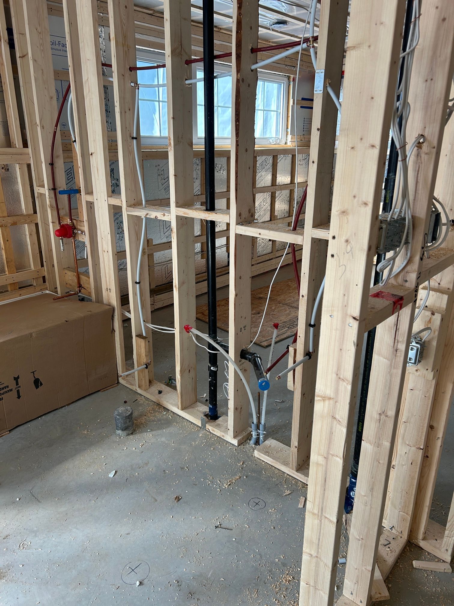 Vue intérieure d'une charpente en bois sur un chantier, avec plomberie et câblage électrique visibles. Fenêtres et sol en béton en arrière-plan.