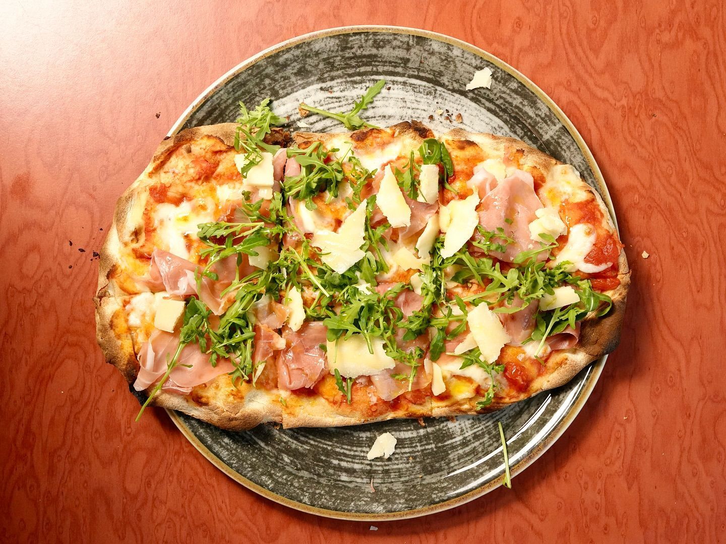 Pizza su un piatto grigio; condita con rucola, prosciutto e formaggio, servita su un tavolo rosso-marrone.