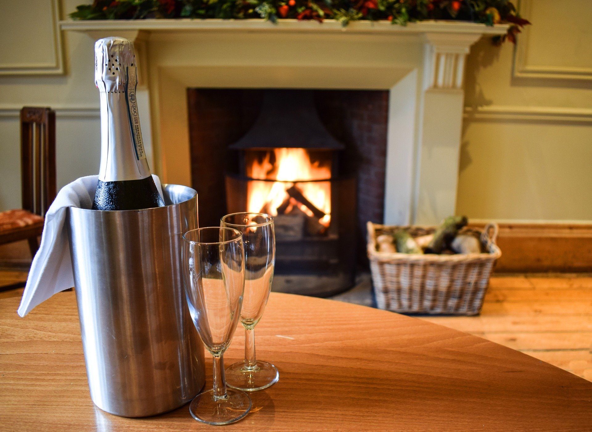 A bottle of champagne in an ice bucket with two glasses on a table in front of a fireplace.