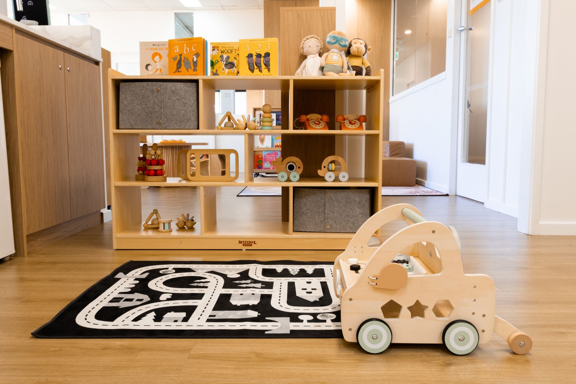 A wooden toy car is sitting on a rug next to a shelf filled with toys.
