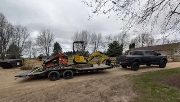 A black pickup truck towing a flatbed trailer loaded with a skid steer and a small yellow excavator on a gravel drive.